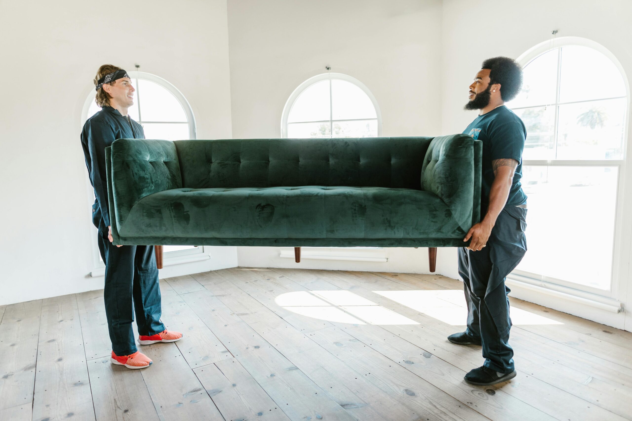 Two men lifting a green sofa in a well-lit room with arched windows.